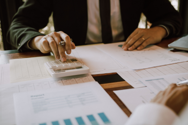 A person in a suit is sitting at a desk full of papers. They are using a calculator while holding a pen in the same hand.